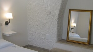 A hotel room with a white textured wall, a bed with a white bedspread, a wall-mounted lamp, a mirror with a gold frame, and a small white shelf. - B&B Gli Archi Country Home (Giurdignano)