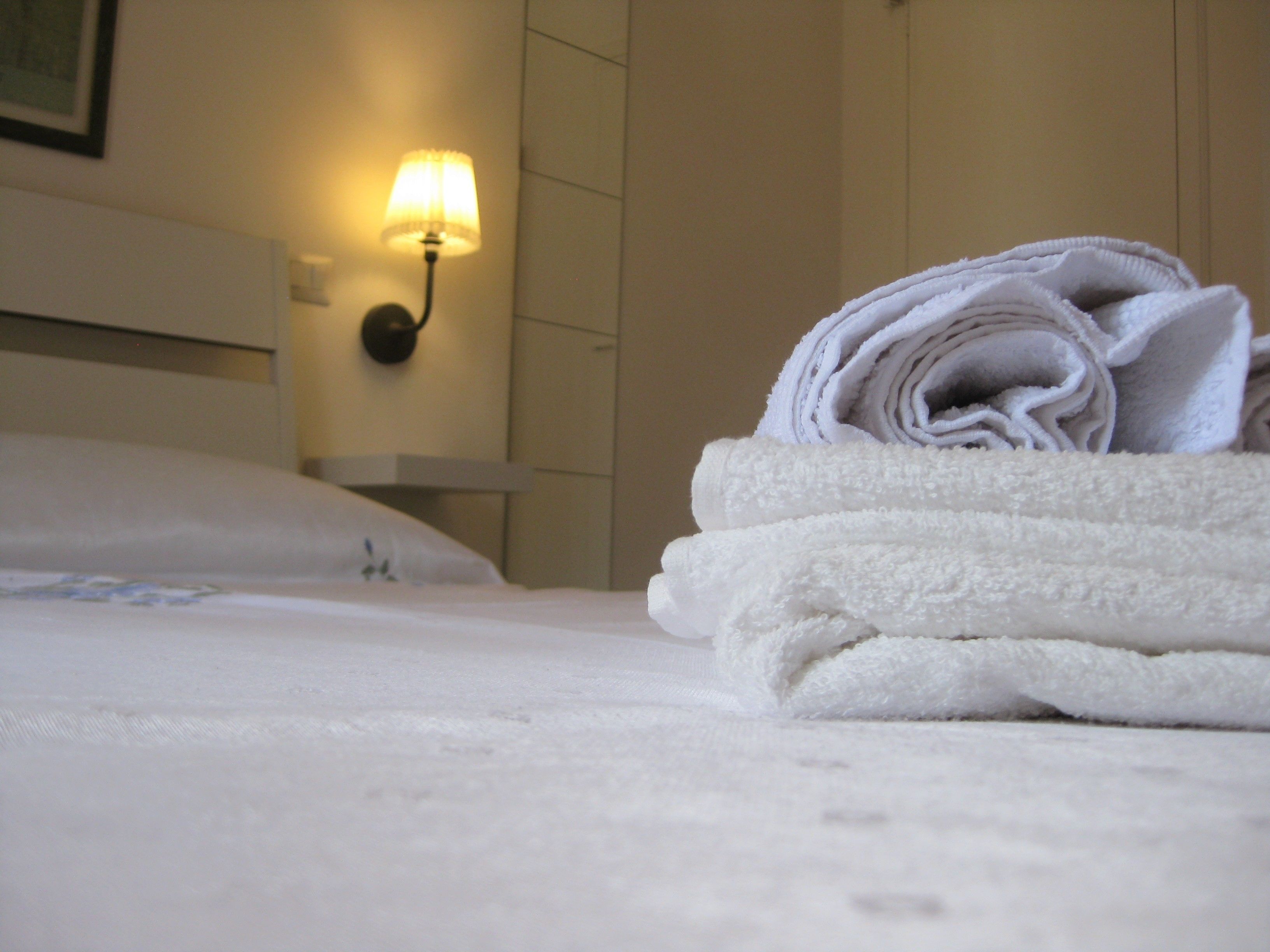 A neatly made bed with a folded white towel on top, a wall-mounted light, and a plain wall in the background.