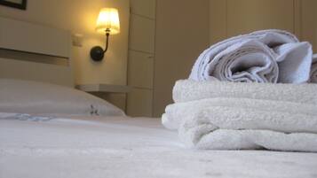 Una cama bien hecha con una toalla blanca doblada encima, una lámpara fijada a la pared y una pared lisa de fondo.