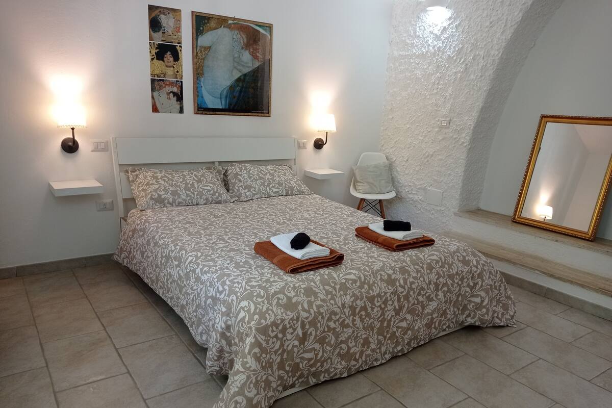 A bedroom with a patterned bedspread, two bedside lamps, a chair, a mirror, and artwork on the wall.