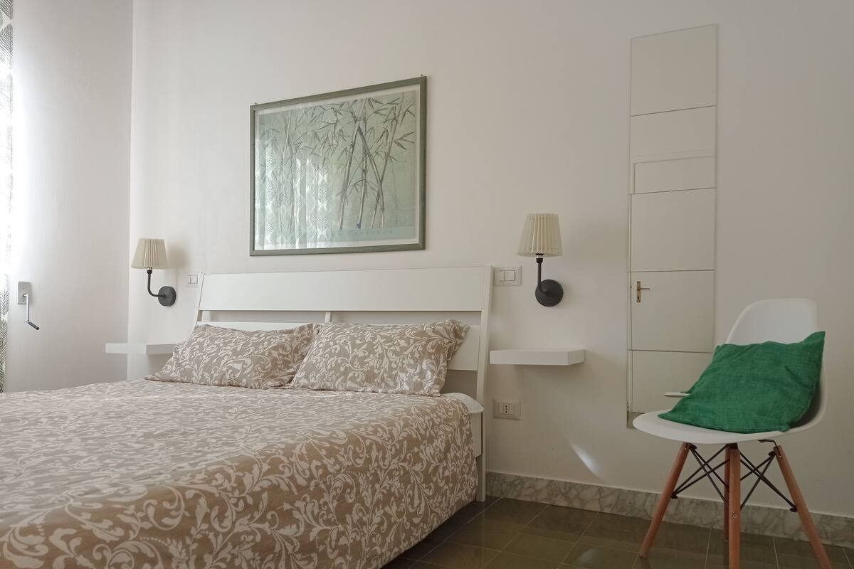 A bedroom with a bed, two wall-mounted lamps, a green chair, and a framed artwork on the wall.
