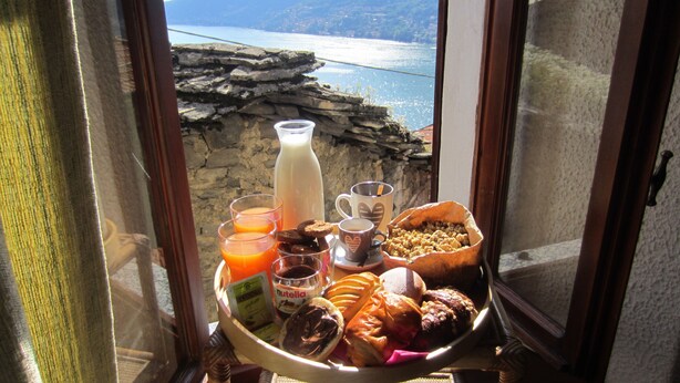 Bandeja de desayuno con café, jugo, galletas y pasteles sobre una mesa junto a una ventana con vista a la costa rocosa.