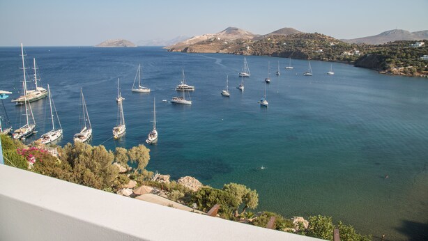 A coastal view with multiple sailboats anchored in calm waters, surrounded by hills and sparse vegetation. - Sunrise Bay. Modern Cycladic House near the beach with a magnificent view. (Leros)