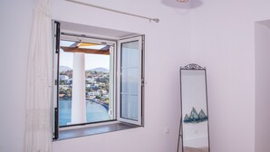 A room with a large window offering a view of a coastal town, a tall mirror, and a neatly made bed with a floral bedspread. - Sunrise Bay. Modern Cycladic House near the beach with a magnificent view. (Leros)