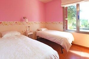 Ein Schlafzimmer mit zwei Betten, einem Nachttisch, einer Lampe und einem Fenster mit Blick auf Grünflächen.