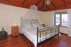 Quarto com cama de dossel, piso de madeira, mesa de cabeceira com abajur e janela com cortinas.