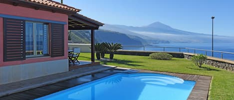 A swimming pool with a wooden deck, a patio area with a table and chairs, and a view of the ocean and mountains.