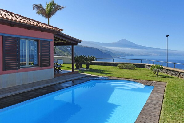 Piscina com deck de madeira, área de estar com mesa e cadeiras, e vista para o oceano e montanhas.