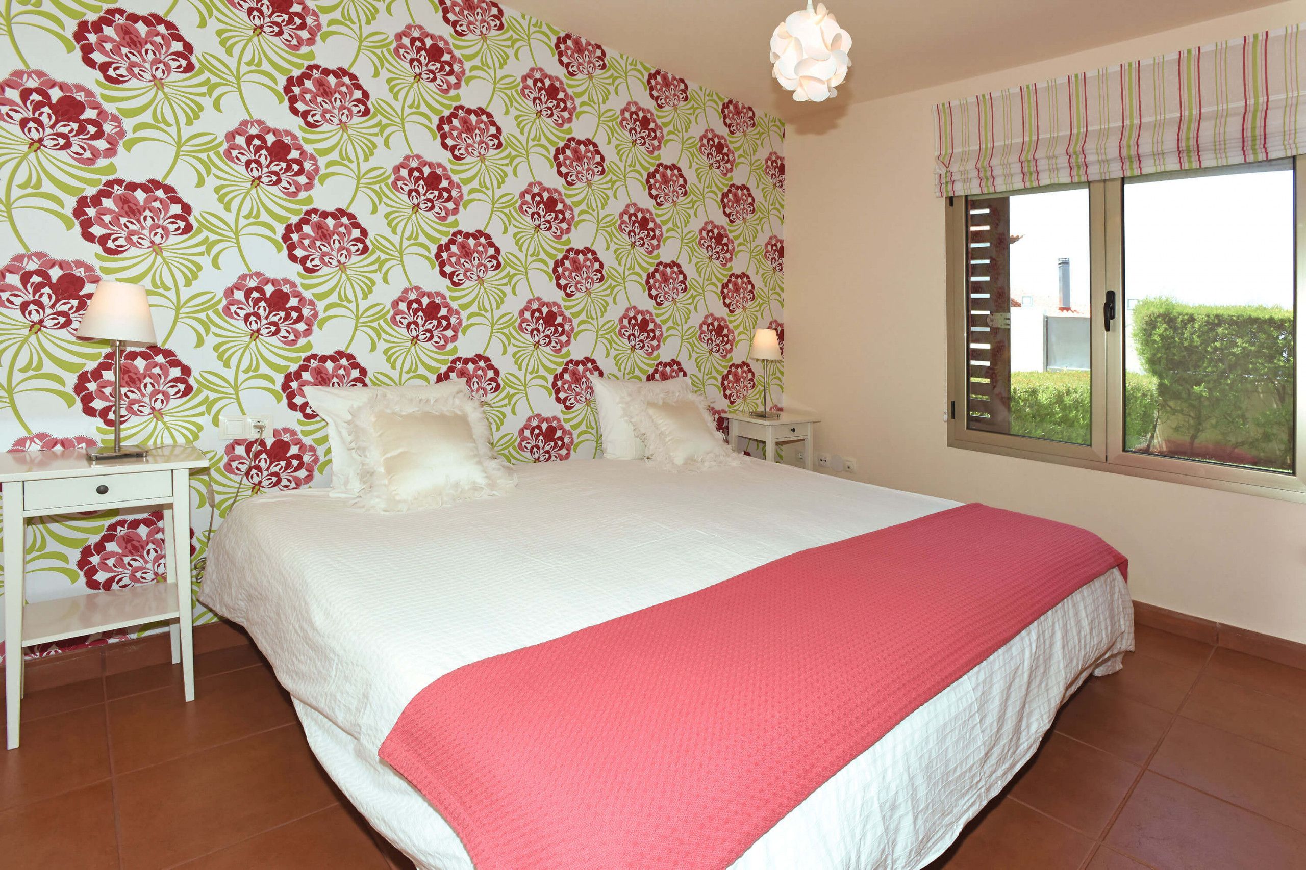 A bedroom with a large bed, floral wallpaper, a window with blinds, and a nightstand with a lamp.