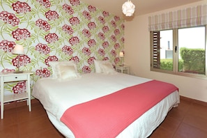 A bedroom with a large bed, floral wallpaper, a window with blinds, and a nightstand with a lamp. - Villa Los Alisios en Tacoronte (Tacoronte)