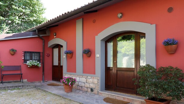 Une maison à l’extérieur rouge, avec des fondations en pierre et une porte en bois. On y trouve des plantes en pot et un banc à l’extérieur.