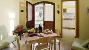 Un coin repas avec une table dressée pour deux personnes, un panier rempli de légumes frais et une vue sur la cuisine par une porte ouverte.