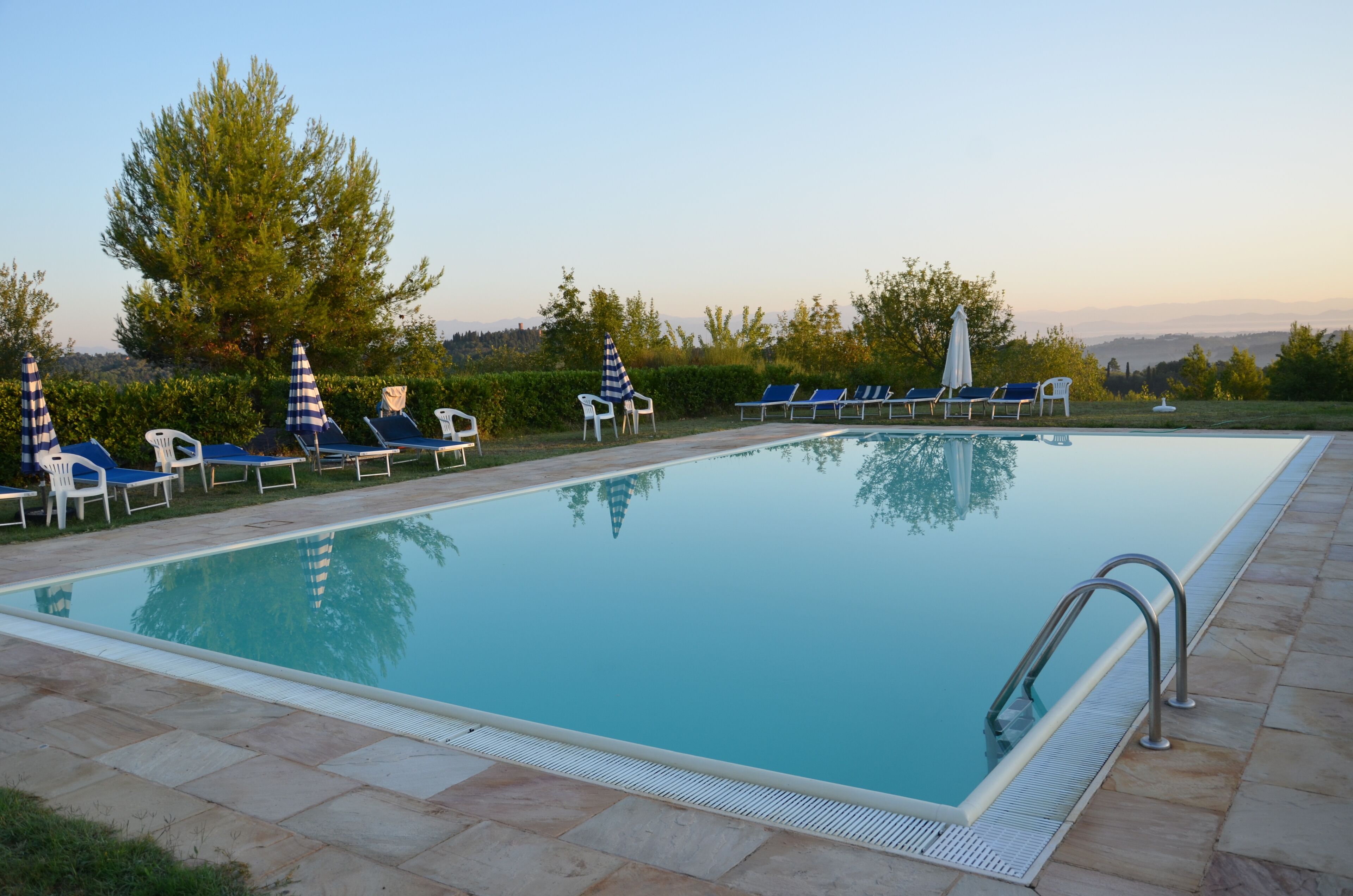 A rectangular swimming pool with lounge chairs and umbrellas around it, set on a stone deck with a view of trees and distant hills.