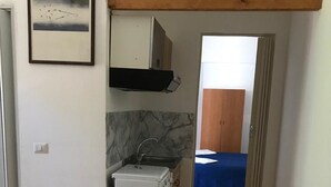 A small kitchenette with a refrigerator, sink, and cabinets. There is a bed with a blue cover in the adjacent room. - Holiday house in Palinuro center (Palinuro)
