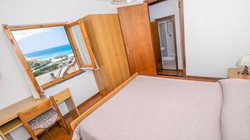 Ein Schlafzimmer mit einem großen Bett, einem Holztisch, einem Stuhl, einem Fenster mit Meerblick und einer Badezimmertür.