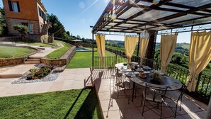Un patio al aire libre con una mesa lista para comer, rodeado por una valla metálica y cortinas, con vistas a un viñedo.
