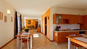 A modern kitchen with wooden cabinets, a dining area with a table and chairs, and a living room with a sofa and a coffee table. - Lake Como - Residence La Collina - Orange (Vercana)