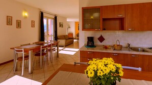 A modern kitchen with wooden cabinets, a dining area with a table and chairs, and a view of the living room through an open door. - Lake Como - Residence La Collina - Orange (Vercana)