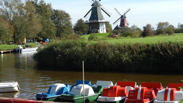 Ein Kanal mit vertäuten Booten, ein Windmühle und eine Wiese mit Bäumen.