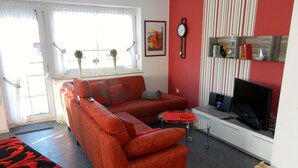 Ein Wohnzimmer mit einer Couch, einem Fernseher, einem kleinen Tisch und einem Fenster mit Vorhängen.