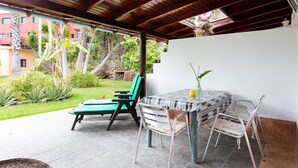 An outdoor patio with a table and chairs, a covered area, and a view of a garden and buildings.