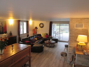 A living room with a sofa, coffee table, and dining table. There is a lamp, a clock, and a view of the outdoors through a sliding glass door. - Grand Lieur Estate (Villeneuve-sur-Vère)
