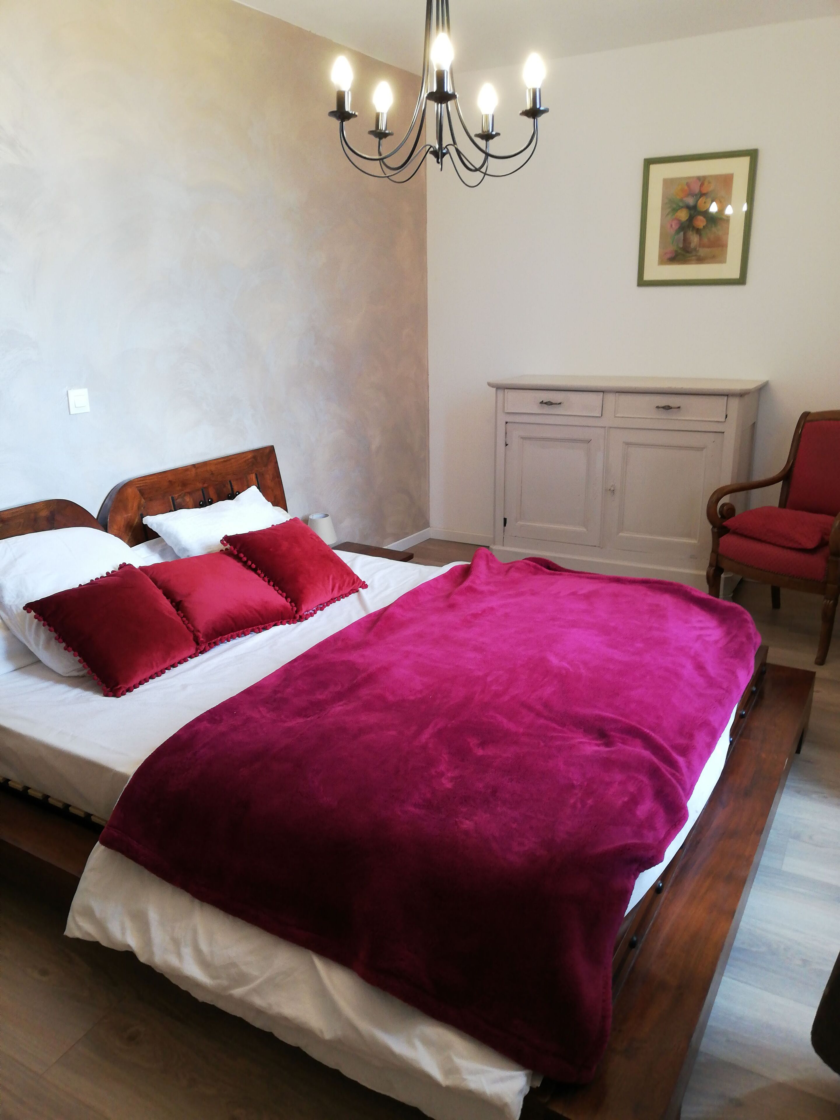 Une chambre avec un grand lit, une tête de lit en bois, une couverture rouge, un lustre, une commode blanche et une chaise rouge.