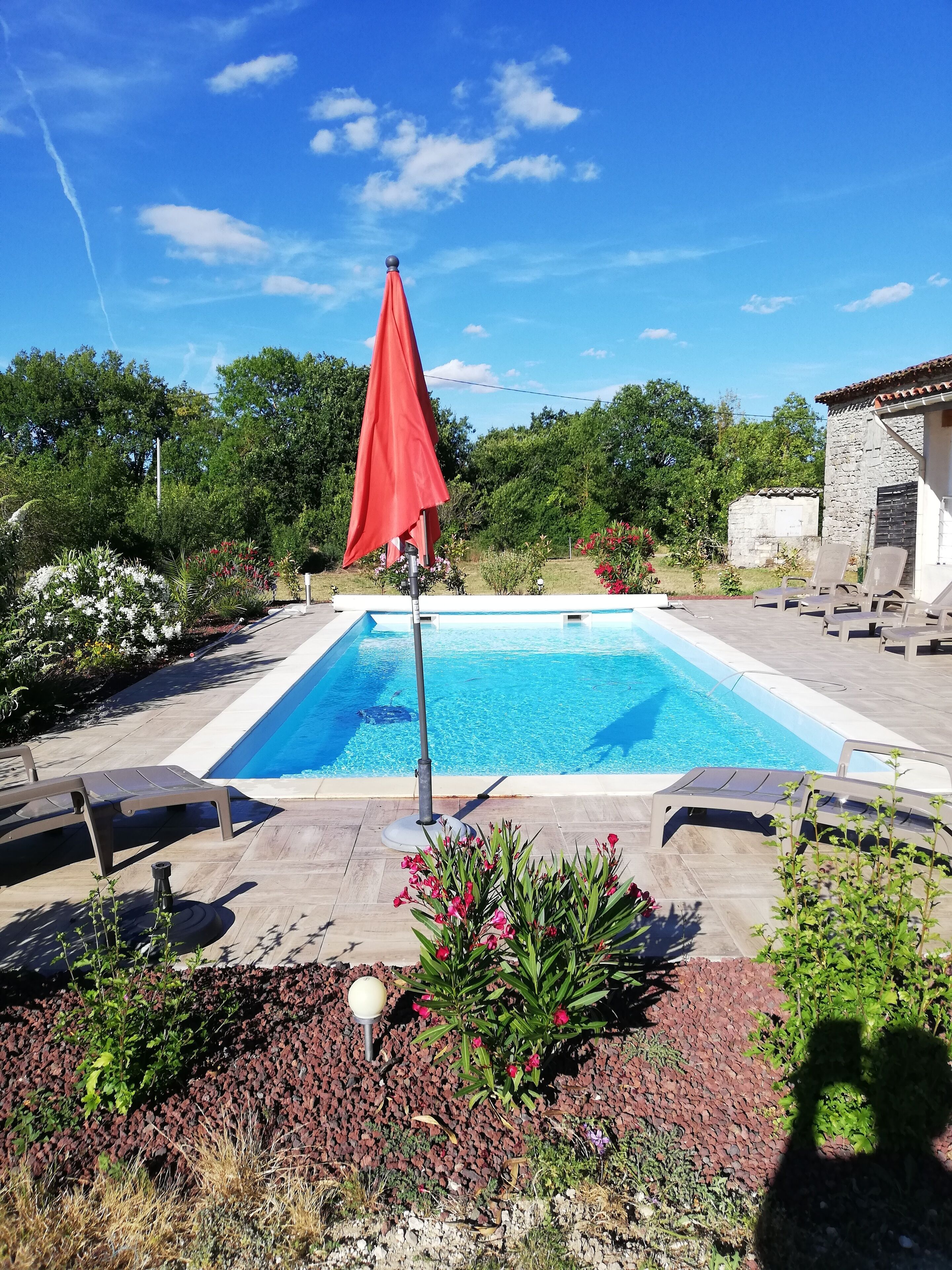 A swimming pool with lounge chairs and a red umbrella, surrounded by landscaped garden beds and a stone building.