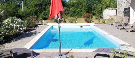 En swimmingpool med liggestole og en rød parasol, omgivet af anlagte bede og en bygning i sten.