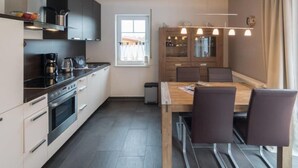 A modern kitchen with a dining area, featuring a wooden table and chairs, a built-in oven, and a window with curtains. - Vacation apartment Meerestraum (Norddeich)