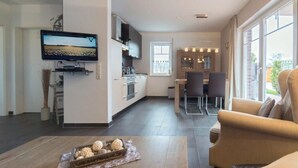 A modern living room with a sofa, a coffee table with decorative items, a TV mounted on the wall, a kitchen area with cabinets and a window, and a dining area with chairs. - Vacation apartment Meerestraum (Norddeich)