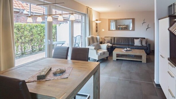 A modern living room with a sofa, a coffee table, and a dining table. There is a large window providing natural light and a view of the outside. - Vacation apartment Meerestraum (Norddeich)