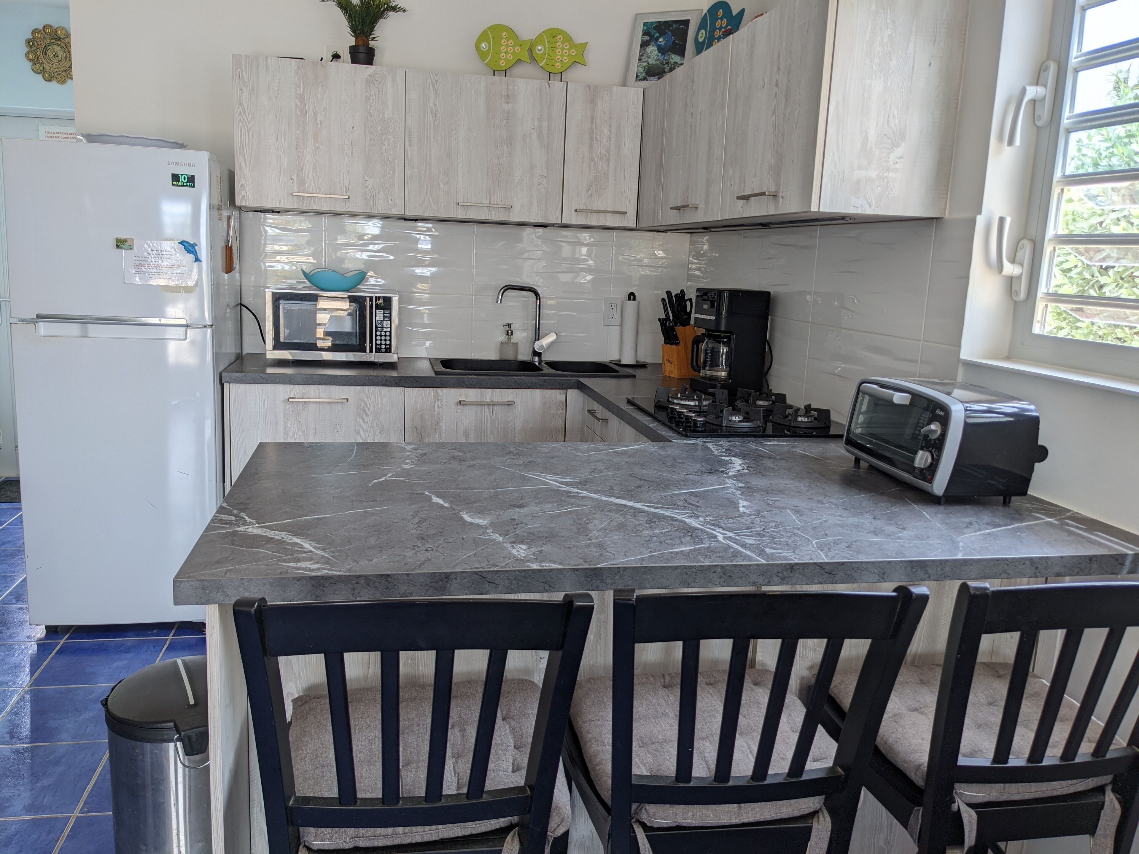 A kitchen with a white refrigerator, a microwave, a toaster, and a sink. There are two chairs and a trash can.