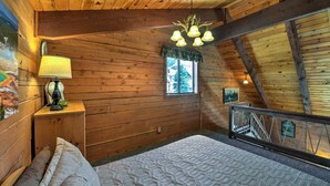 Una camera accogliente con pareti in legno, un letto con lenzuola decorate, un comodino con una lampada, una finestra vista alberi e una ringhiera da balcone.