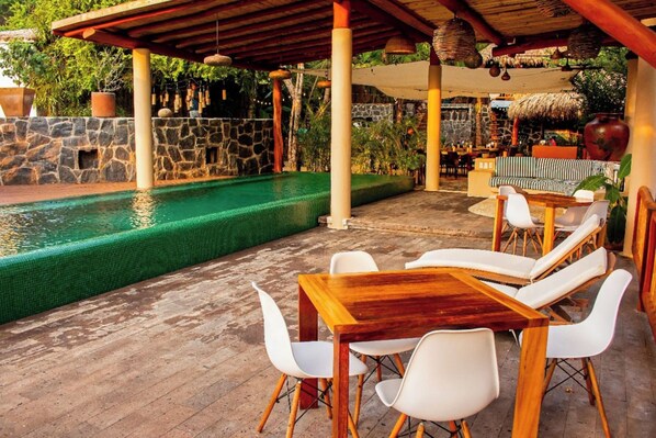 Un espace extérieur avec une piscine, une table et des chaises en bois, un mur en pierre et une structure dotée d’un toit de chaume.
