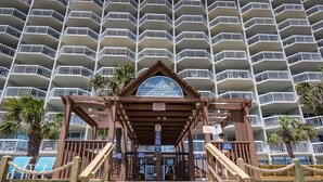 Una entrada de madera a la playa con un cartel anunciando Oceanfront Condominiums, mostrando una vista de un edificio de gran altura con balcones.