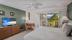 Un dormitorio con cama, escritorio, silla, televisión y balcón con vistas a palmeras y una playa.