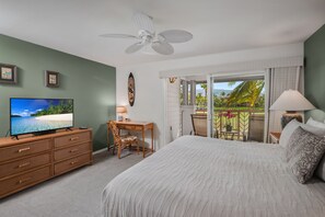 Een slaapkamer met een bed, een bureau, een stoel, een televisie en een balkon met uitzicht op palmbomen en een strand.