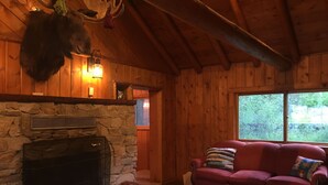 L’intérieur chaleureux d’un chalet avec une cheminée en pierre, un canapé rouge et une grande fenêtre donnant sur un paysage verdoyant.