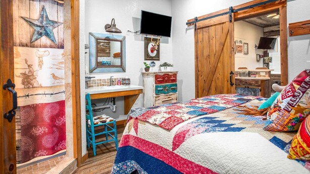 A rustic bedroom with a bed, a dresser, a desk, and a TV. - Lonesome Dove Guesthouse (Bandera)