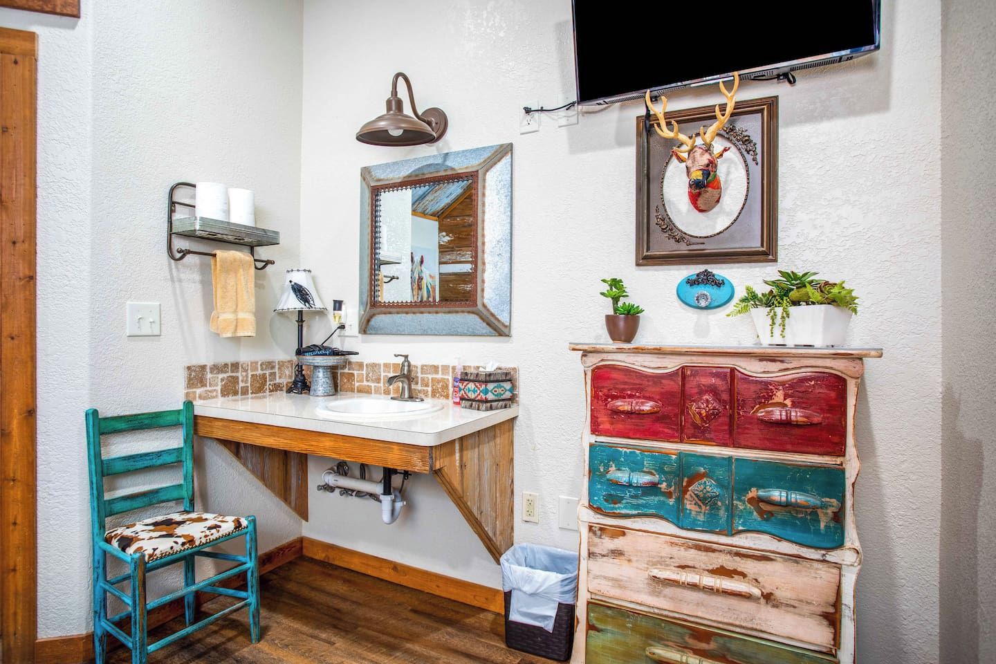 A bathroom with a wooden vanity, a sink, a mirror, a towel rack, a wall-mounted lamp, a television, and a decorative deer head.