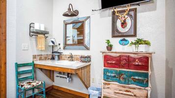 A bathroom with a wooden vanity, a sink, a mirror, a towel rack, a wall-mounted lamp, a television, and a decorative deer head.