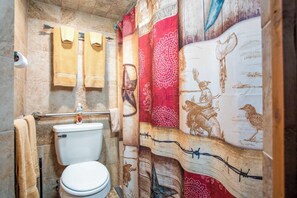 A bathroom with a toilet, a towel rack with towels, and a shower curtain with a patterned design. - Lonesome Dove | 1BR Cabin Near 11th Street Bar (Bandera)