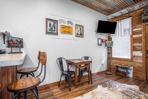 A room with a wooden table, two chairs, a mounted television, and wall-mounted artwork. - Lonesome Dove | 1BR Cabin Near 11th Street Bar (Bandera)