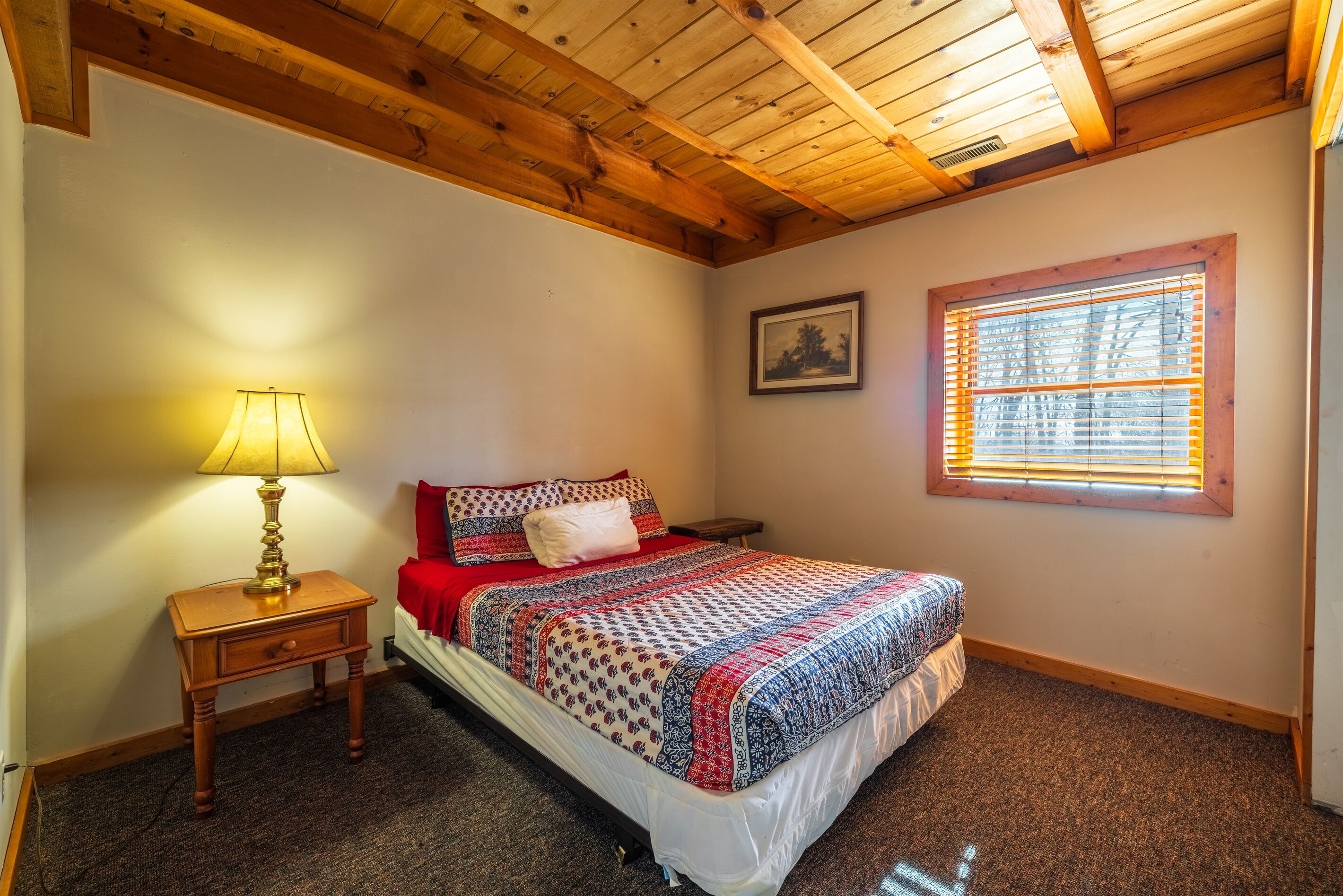 A bedroom with a wooden ceiling, a bed with patterned bedding, a nightstand with a lamp, a window with blinds, and a framed picture on the wall.