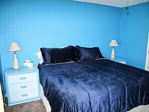 Une chambre avec un mur bleu, un lit recouvert d’une couvre-lit bleu foncé, deux tables de chevet avec des lampes et une petite table de nuit blanche avec des tiroirs.