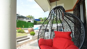 Uma cadeira suspensa com almofadas vermelhas em área externa, com vista para uma casa e piscina.