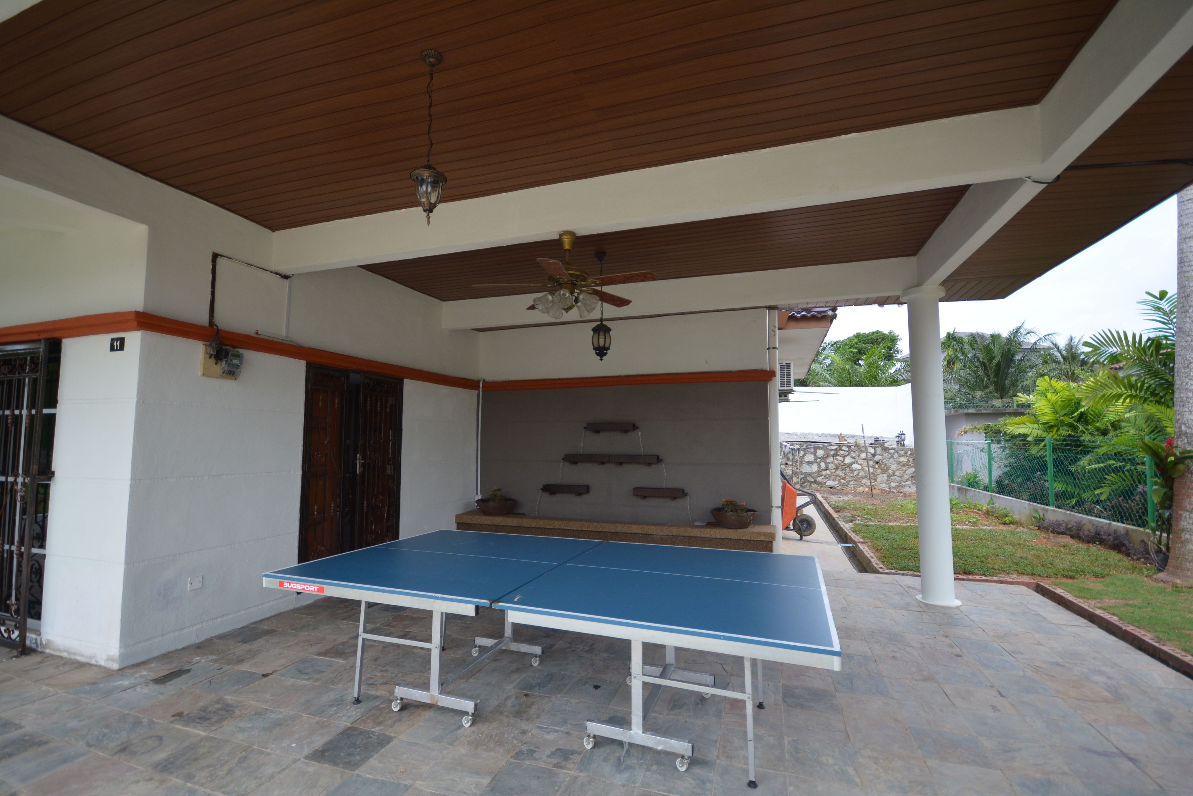 A covered outdoor area with a ping pong table, a ceiling fan, and a stone wall with plants.