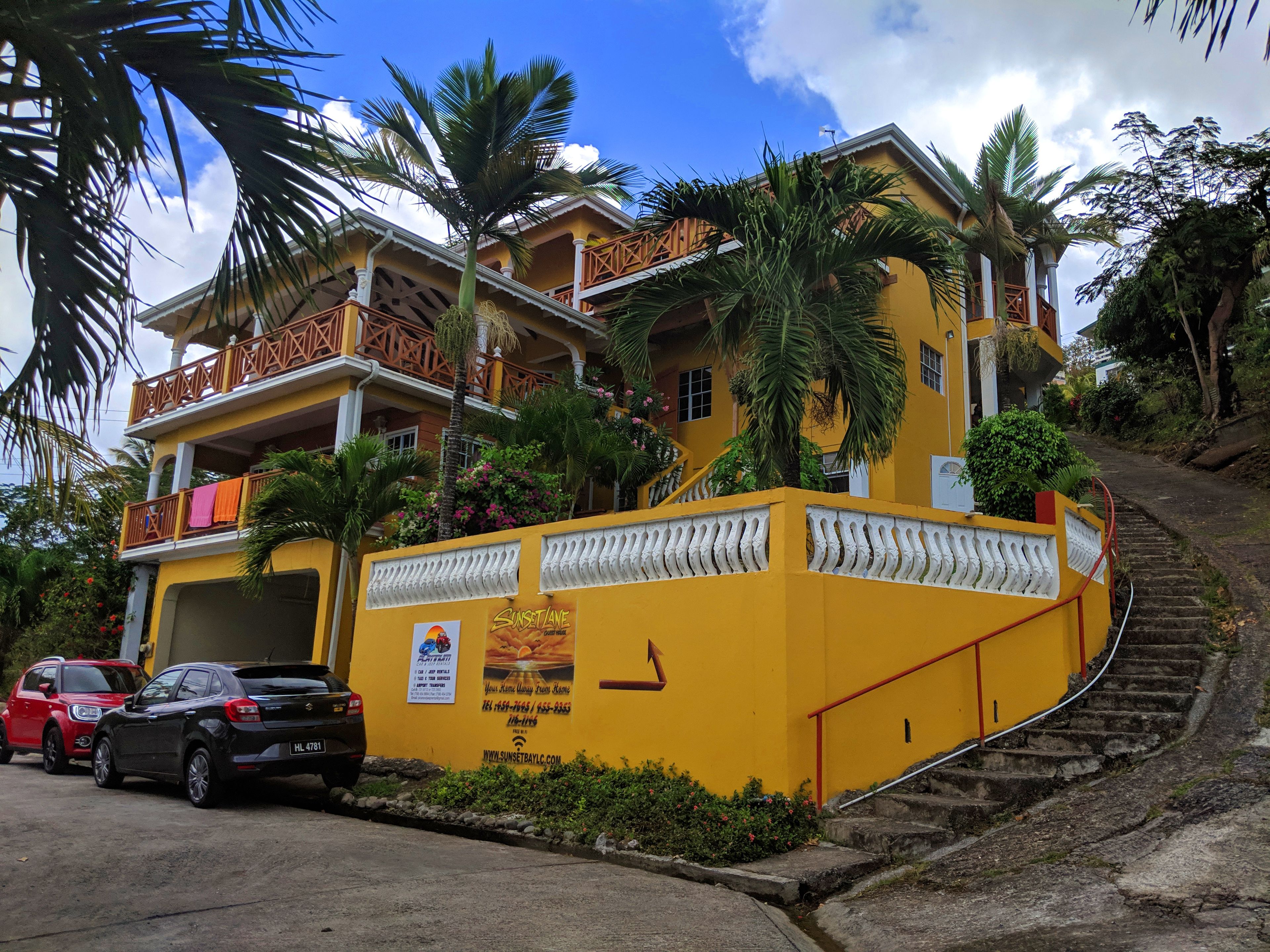 Um edifício amarelo com uma escadaria que leva a uma varanda, rodeado por palmeiras e céu claro.