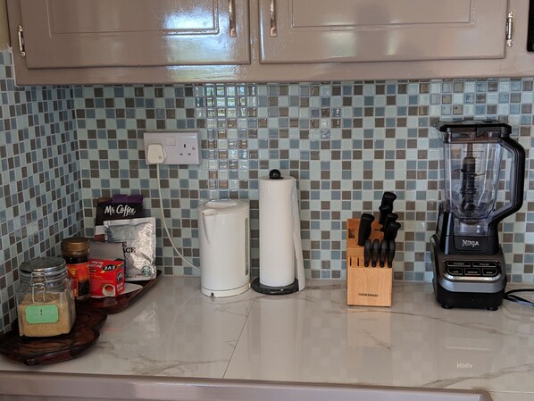 A kitchen counter with a blender, a knife block, a jar of sugar, a box of coffee, and a paper towel holder.